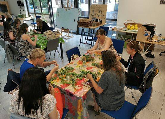 grupa ludzi siedzących przy kilku stołach. Uczestnicy warztatu zielarskiego wykonują preparaty octowe. Na stołach rośliny. fot. JK