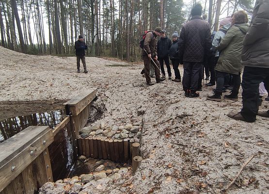 Na zdjęciu na pierwszym planie drewniana zastawka blokująca odpływ wody z rowu, w tle uczestnicy spaceru.