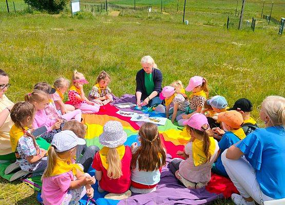 Grupa dzieci siedzą w okręgu na kolorowe chuście, rozłożonej na łące