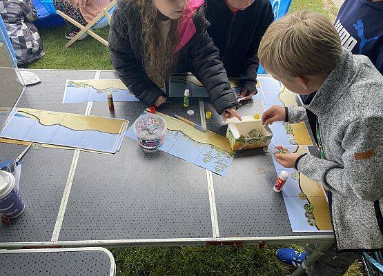 Dzieci pracujące przy stole nad planszami edukacyjnymi z ekosystemem nadmorskim, używające kleju i naklejek podczas warsztatów przyrodniczych.