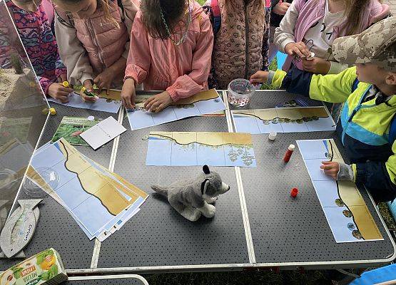 Dzieci pracujące przy stole nad planszami edukacyjnymi z ekosystemem nadmorskim, używające kleju i naklejek podczas warsztatów przyrodniczych.