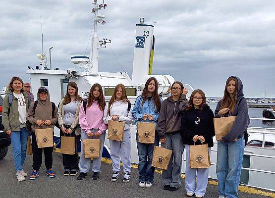 Uczniowie z opiekunami pozują do zdjęcia na tle kutra edukacyjnego. Każdy trzyma torbę z nagrodami