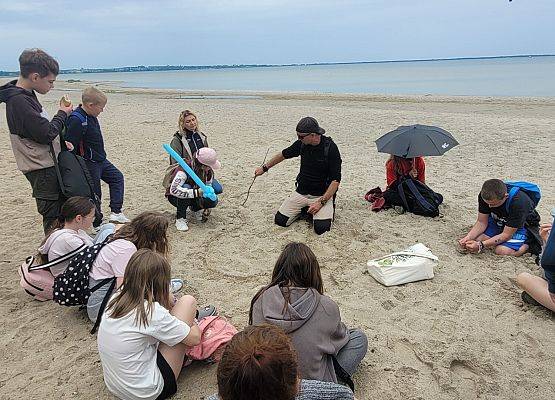 Uczniowie siedzą na plaży w kółku. Instruktor rysuje na piasku i omawia proces powstania mierzei