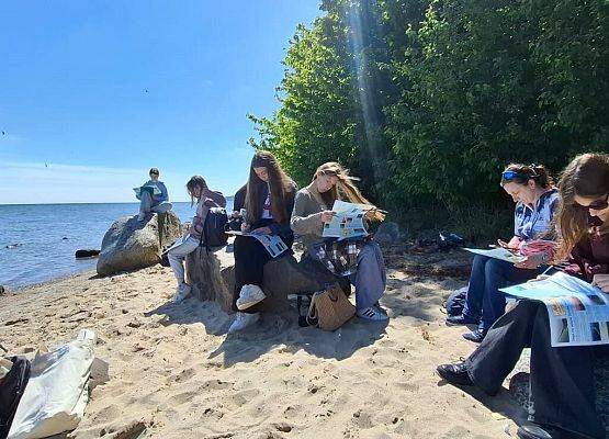 Słoneczny dzień. Uczniowie (ok 10 os) nad brzegiem zatoki, siedzą na głazach i rozwiązują zadania nakacie pracy.