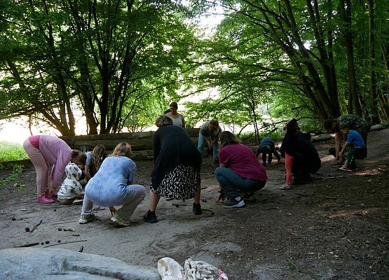 Zdjęcie przedstawia grupę osób – dorosłych i dzieci – pochylających się ku ziemi w lesie,  biorących udział w warsztatach ruchowych na świeżym powietrzu. Uczestnicy są skupieni, wykonują synchroniczny ruch, jakby coś zbierali lub dotykali ziemi.