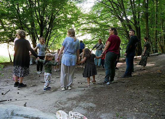 Zdjęcie przedstawia grupę ludzi trzymających się za ręce i stojących w kręgu w lesie, na leśnej polanie otoczonej drzewami. W kręgu uczestniczą zarówno dorośli, jak i dzieci, co nadaje scenie rodzinny i integracyjny charakter. W tle widoczne są