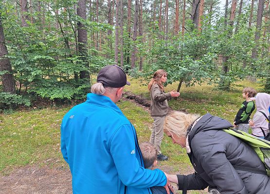 Przewodniczka w lesie gestykuluje, opowiadając zgromadzonej grupie dorosłych i dzieci podczas spaceru edukacyjnego wśród wysokich sosen.