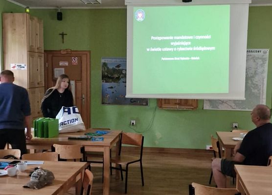 Spotkanie z Państwową Strażą Rybacką. Fot. Ł.Pepliński Zdjęcie przedstawia salę z zielonymi ścianami i drewnianą podłogą, w której kilka osób siedzi przy stołach. Na ekranie wyświetlana jest prezentacja w języku polskim.