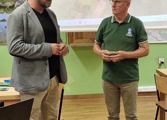 Spotkanie z Państwową Strażą Rybacką. Fot. Ł.Pepliński Dwaj mężczyźni rozmawiający na sali, w tle ekran multimedialny na którym prezentowany jest zdjęcie jeziora z lotu ptaka.