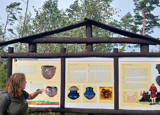 Spacer z okazji Dnia Parków Krajobrazowych „W poszukiwaniu archeofitów” fot. Karolina Rozmarynowska Przewodniczka przy tablicy obrazującej różne kultury osadnicze okolic leśna: łużycką, pomorską i wielbarską. Na planszy różne typy glinianych popielnic.