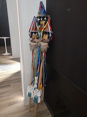 W rogu nowoczesnego wnętrza, przy ciemnej ścianie, oparte są trzy pary tradycyjnych ludowych instrumentów zwanych "diabelskimi skrzypcami". Instrumenty mają ozdobne, kolorowe dekoracje: trzy drewniane głowy z groźnymi, wyrazistymi