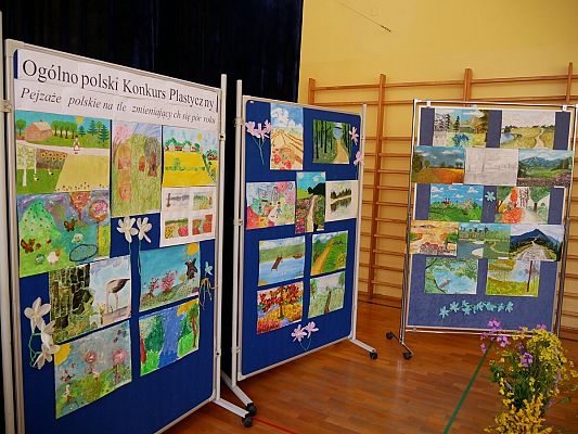 Wnętrze budynku z drewnianymi drabinkami powieszonymi na ścianach i z drewniana lakierowana podłogą. Na zdjęciu widoczna jest wystawa prac plastycznych, złożona z trzech dużych, granatowych, stojących pionowo tablic. Na każdej tablicy znajdują