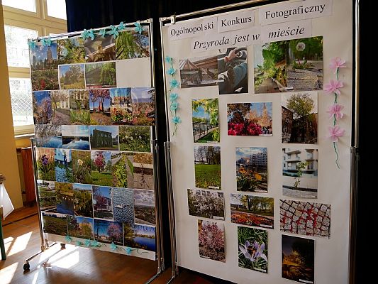Wnętrze budynku z dużymi oknami, z założonymi białymi siatkami ogrodzeniowymi na framugach od zewnątrz. Na zdjęciu widoczna jest wystawa fotograficzna, złożona z dwóch dużych, białych, stojących pionowo tablic. Na każdej tablicy znajdują się