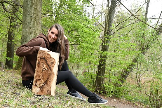 Sylwia z dziełem, fot. A. Janczyszyn Kobieta siedząca na trawie, przed sobą trzyma deskę z wypalonym portretm lwa. W tle las