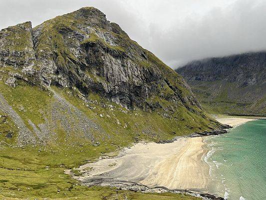 Góry skaliste, z zieloną trawą, po prawej stronie brzeg wody. W oddali chmury.